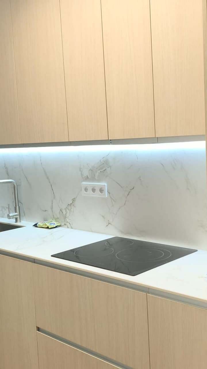 Vídeo de una reforma de cocina: muebles altos de madera clara, encimera blanca con placa y fregadero, frontal veteado con luz bajo los muebles y muebles bajos azules